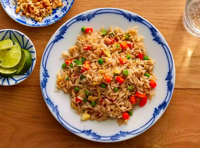 A bird’s-eye view of fried rice with carrots and peas, served on a blue and white plate, with limes and peanuts in small bowls beside it.