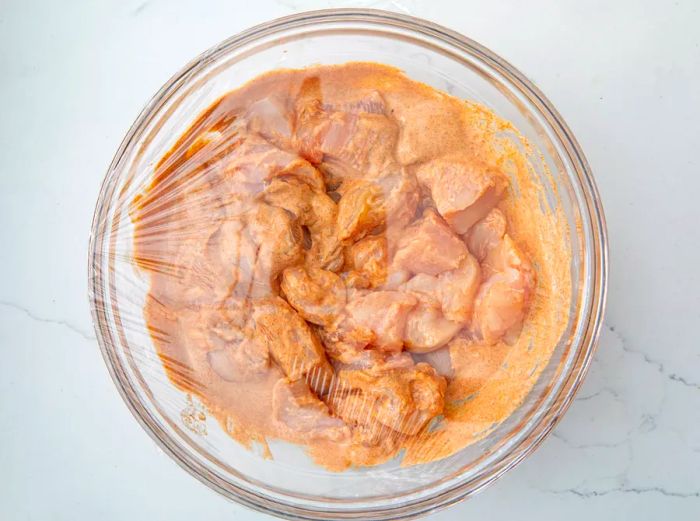 Chunks of chicken coated and soaking in the Tikka Masala marinade in a bowl.