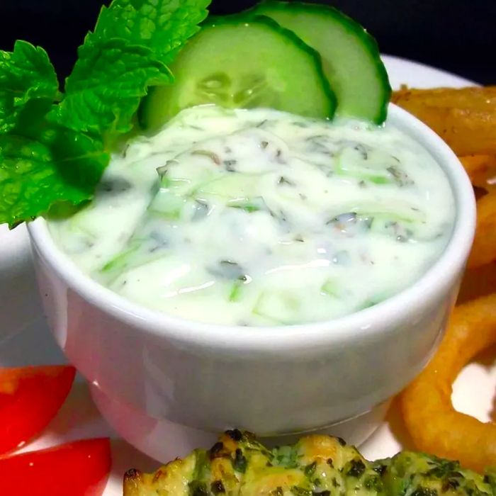 A close-up view of a small dish of cucumber raita, with fresh cucumbers inside.