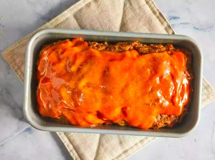 An overhead shot of a meatloaf coated with sauce, resting in a baking pan.