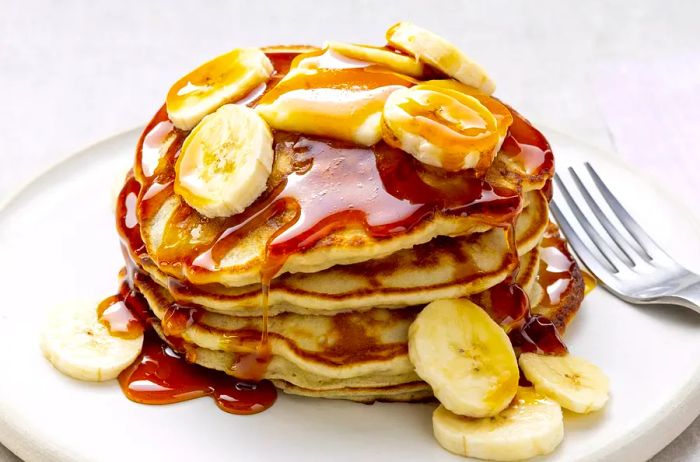 A close-up shot of a scrumptious stack of banana pancakes topped with fresh banana slices, syrup, and a dollop of melting butter.