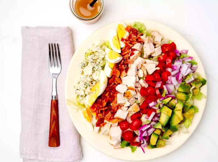 A beautiful Cobb salad topped with fresh ingredients, served on a white plate alongside a fork, napkin, and dressing.