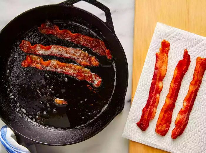 An overhead view of bacon sizzling in a cast iron skillet, with three crispy strips resting on a paper towel.