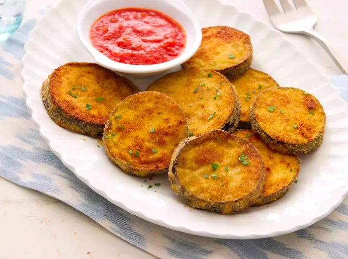 A plate of golden fried eggplant served alongside a small dish of tomato sauce.