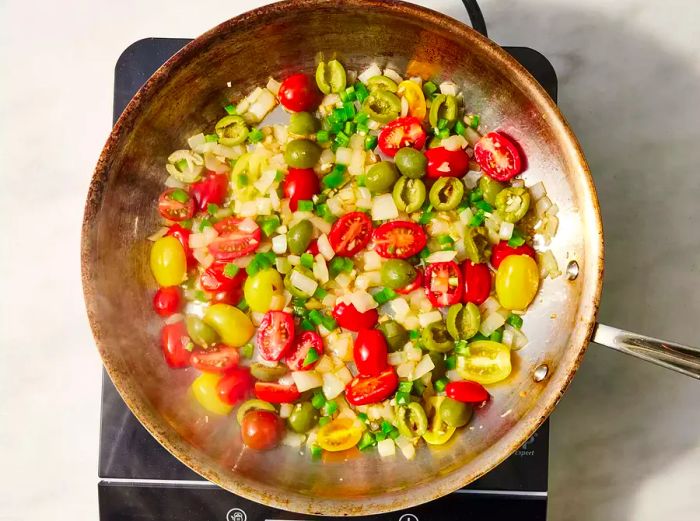 Onions, tomatoes, olives, and jalapeños cooking together in a pan on the stovetop