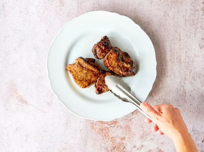 A cooked steak resting on a white plate with tongs on a marbled countertop.