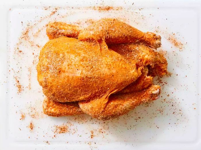 A white cutting board with a chicken coated in spice rub