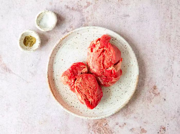 Raw steak seasoned with salt and pepper, placed on a plate, with seasonings on the countertop.