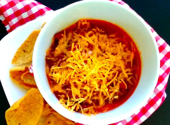 Close-up of Homemade Chili topped with melted shredded cheese in a white bowl, accompanied by a side of crunchy chips