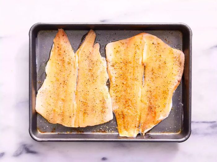 A baking sheet with two flounder fillets drizzled with olive oil, sprinkled with garlic salt, and seasoned with pepper