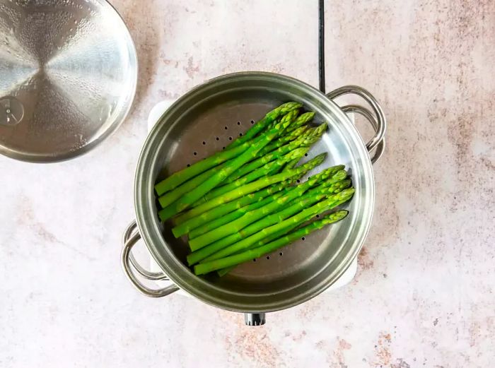 Steaming fresh green asparagus in a metal steamer over a pot on the stove.