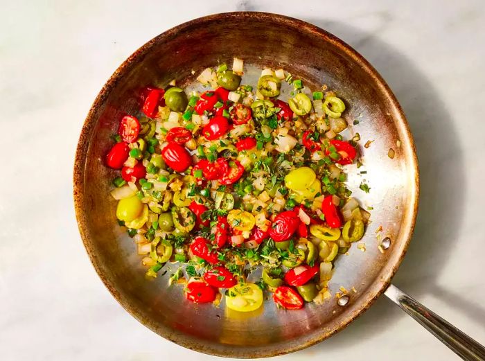 Onions, tomatoes, olives, and jalapeño cooking together in a pan, seasoned with oregano