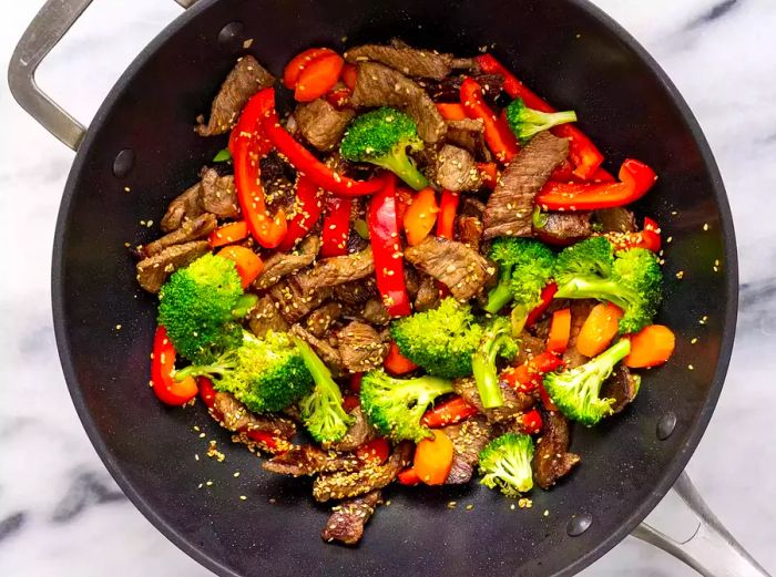 Aerial view of chopped vegetables, beef, and sesame seeds cooking together in a wok.