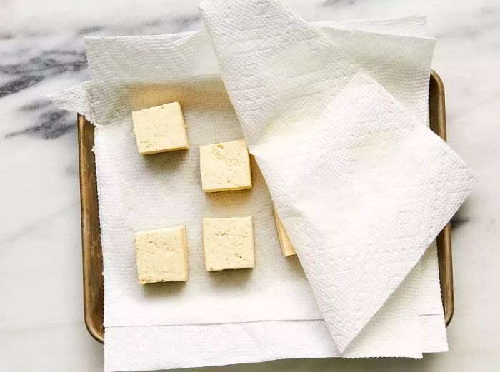 Tofu slices placed between paper towels on a baking sheet