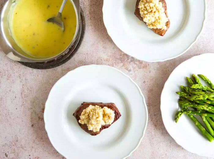 Plates with cooked steak, asparagus, butter sauce, and feta cheese on the countertop.