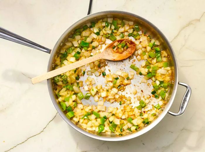 Onion, bell pepper, jalapeño, and garlic being sautéed in a pan with a wooden spoon