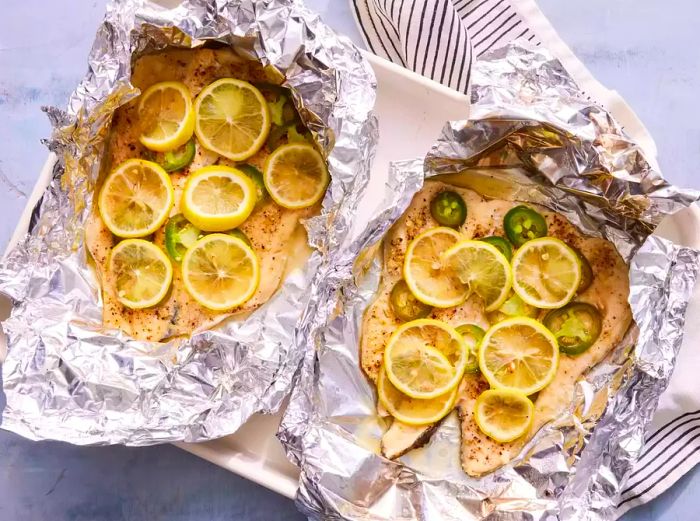 A serving platter with two opened foil packets containing baked trout fillets, garnished with lemon slices and jalapeno slices