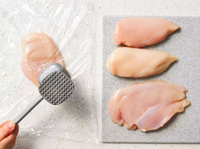 A hand using a mallet to flatten a chicken breast on a grey cutting board, with three additional chicken breasts beside it.