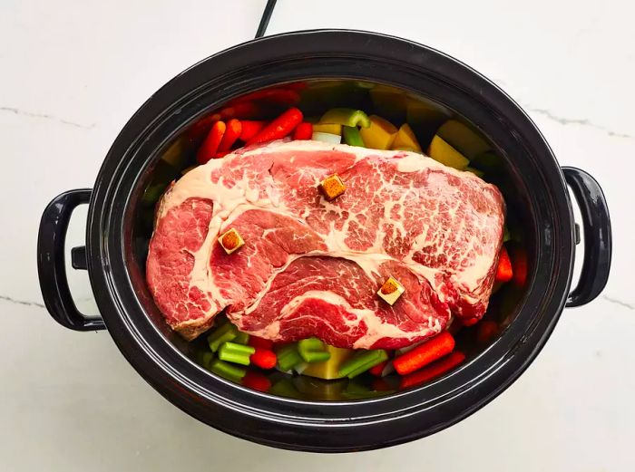 Pot roast in the crockpot resting atop vegetables with bouillon cubes sprinkled on top.