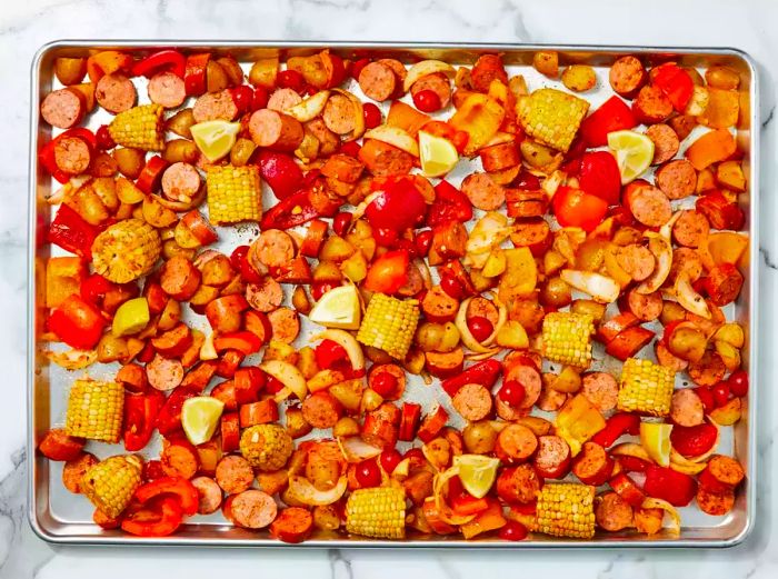 A large baking sheet is covered with seasoned sausage, corn, bell peppers, onions, and cherry tomatoes.