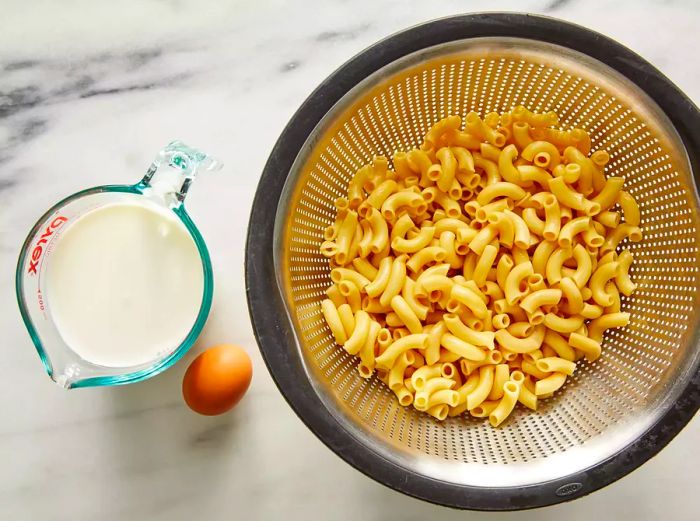 A top-down view of the boiled macaroni in a strainer, with milk and an egg nearby, ready for making mac and cheese.