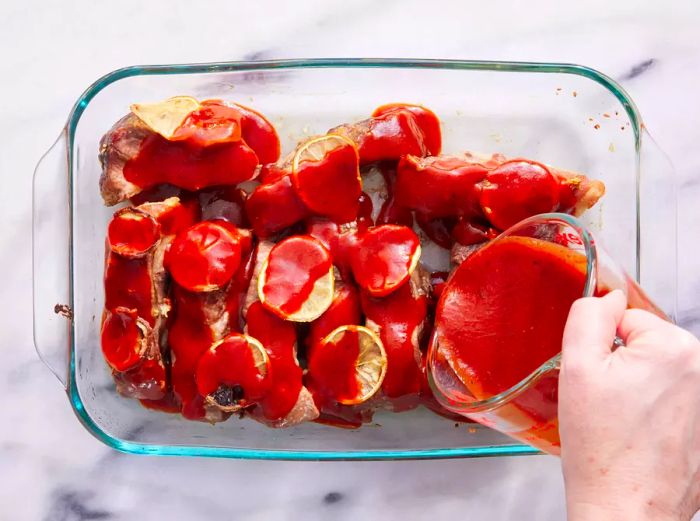 A hand drizzling barbecue sauce over roasted ribs in a baking dish