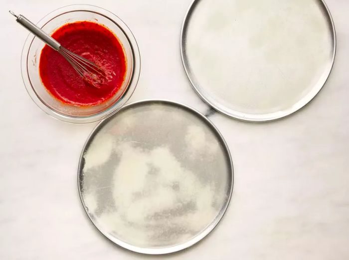 A top-down view of pizza sauce in a bowl alongside two pizza pans.