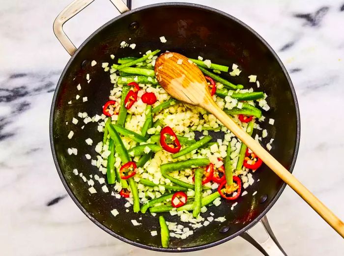 A wok with a stir-fry of onion, red chilies, and bell pepper being mixed with a wooden spoon