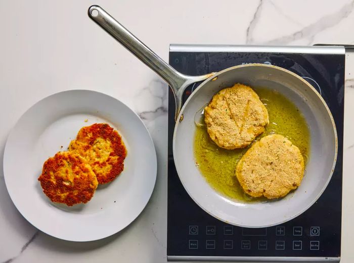 A pan with two chicken burger patties sizzling in olive oil, beside a plate with two fully cooked patties.