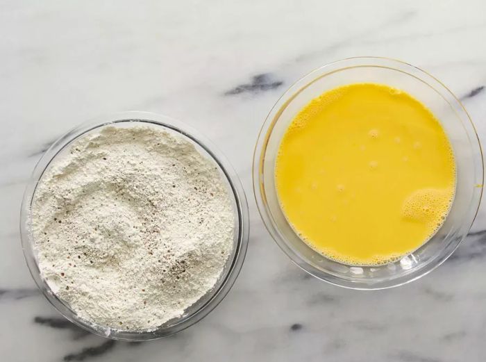 An overhead view of flour dredge and whisked eggs in two separate glass bowls