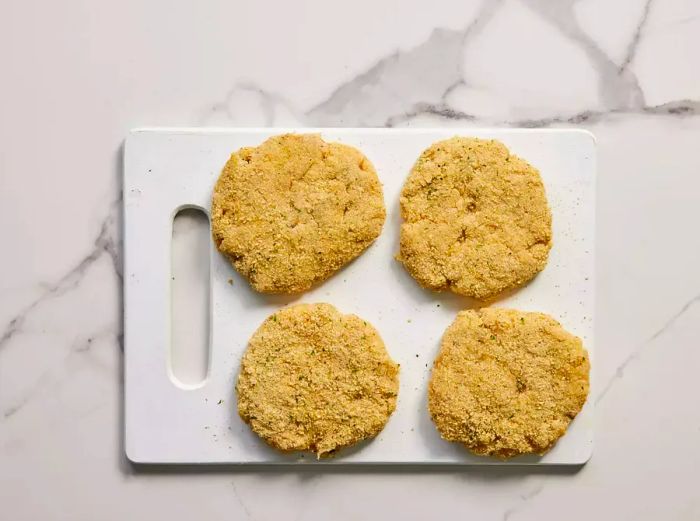 A cutting board with four breaded chicken burger patties ready to cook.