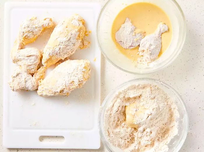 A cutting board with chicken pieces coated in flour, beside a bowl of chicken dipped in beer batter, and another bowl of chicken in the seasoned flour mix