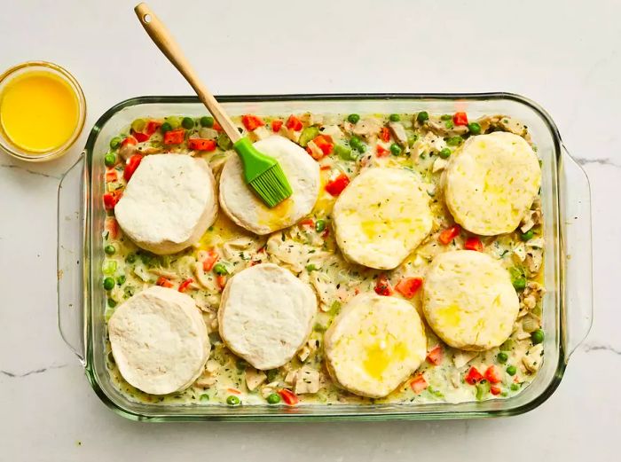 A top-down view of the chicken pot pie casserole topped with raw biscuits being brushed with the egg wash.