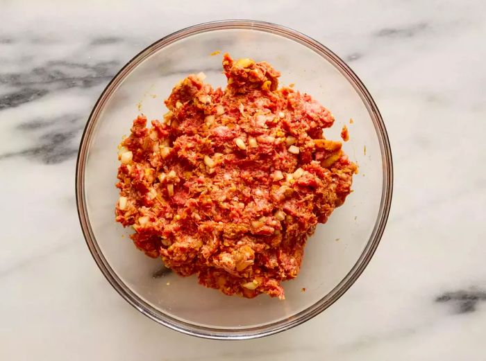 A large bowl filled with ground beef, croutons, sweet onion, egg, garlic, Worcestershire sauce, mustard, red pepper flakes, and Cajun seasoning all blended together