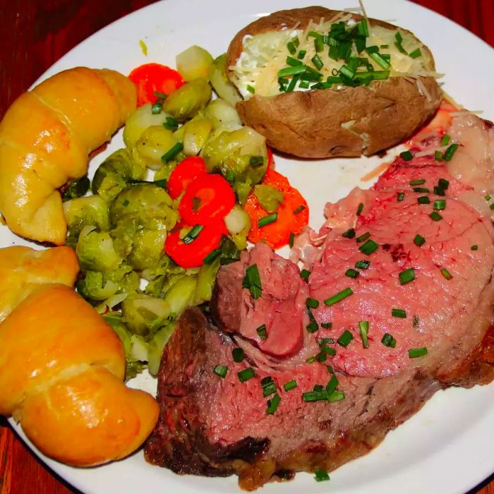 A close-up shot of a slice of Restaurant-Style Prime Rib Roast served with vegetables, baked potato, and dinner rolls on a white plate.