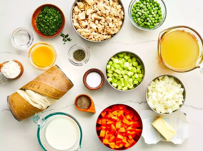 A bird's-eye view of all the ingredients for mom's iconic chicken pot pie with a biscuit topping arranged on a marble counter.