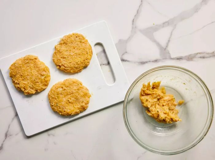 A cutting board with three chicken burger patties next to a bowl of seasoned ground chicken mixture.