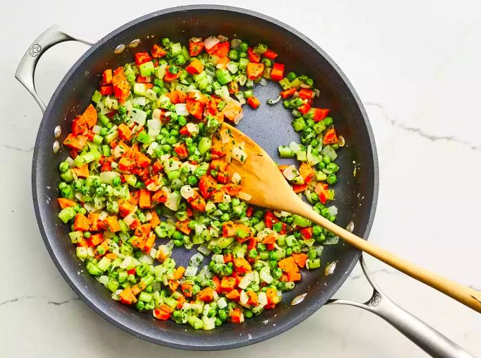 A top-down view of peas, carrots, onions, celery, parsley, and flour sizzling together in a skillet.