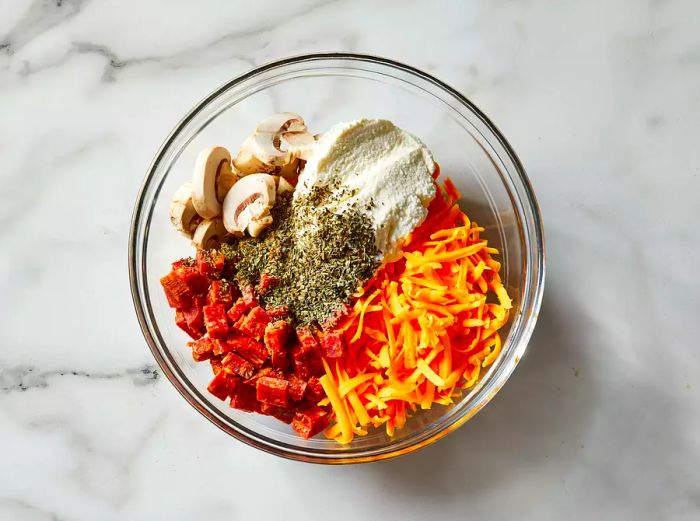 A top-down shot of a mixture of Cheddar cheese, ricotta cheese, pepperoni, mushrooms, and basil in a glass bowl