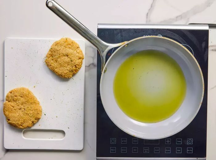 A skillet with olive oil placed next to a cutting board holding two chicken burger patties.