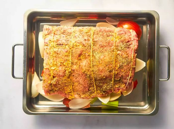 Prime rib, tied and seasoned, resting on a bed of vegetables on a baking sheet.