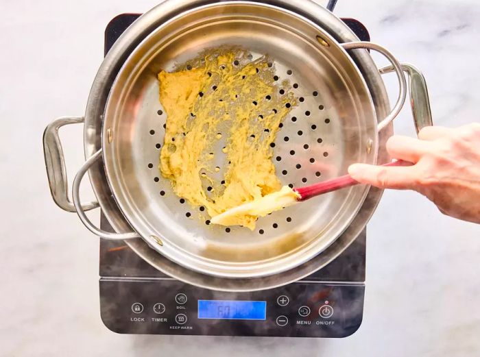 A spatula pressing spaetzle batter through the holes of a strainer into a pot of simmering water