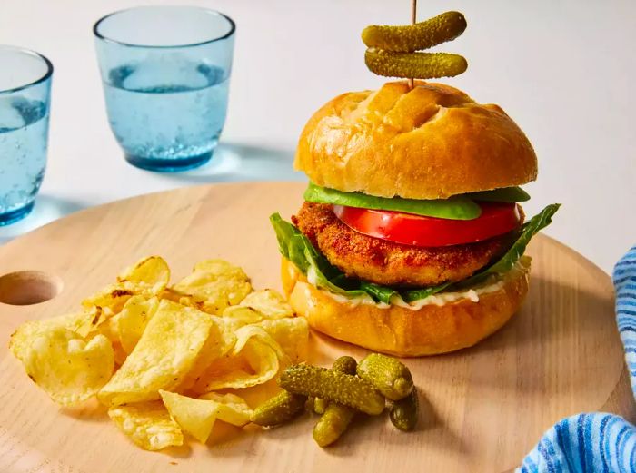 A chicken burger placed on a bun with lettuce, tomato, mayo, and avocado, accompanied by a side of potato chips.