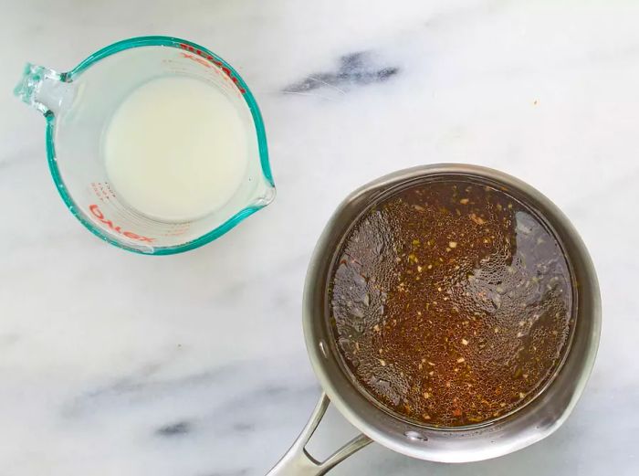 Overhead view of marinade being added to a saucepan, with cornstarch and water in a measuring cup ready to mix.