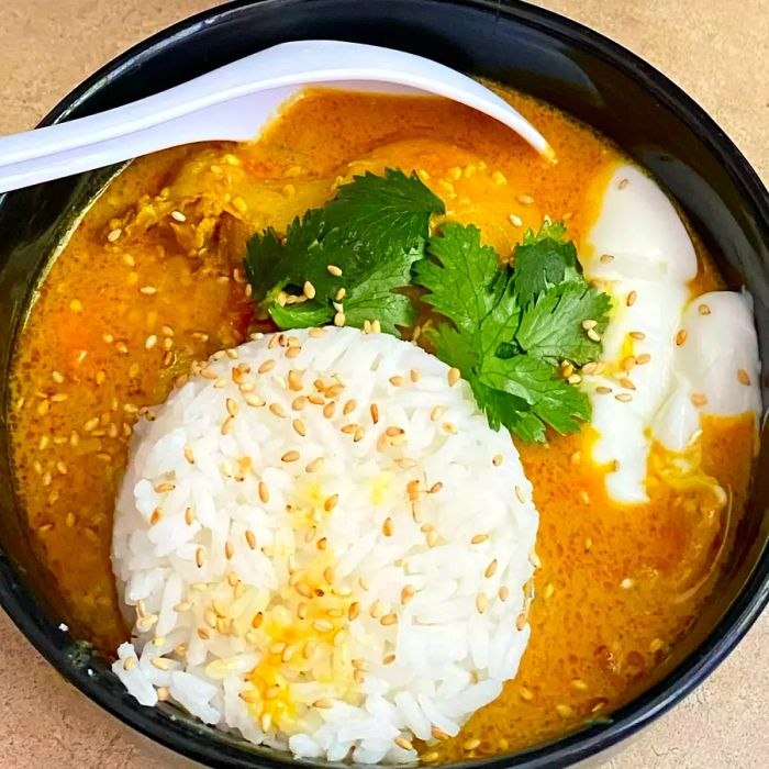 Close-up shot of Peanut Curry Chicken in a black bowl with white rice, fresh herbs, and garnished with sesame seeds.