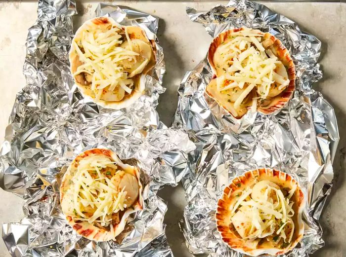 Four scallop shells filled with mushrooms, scallops, and grated cheese, resting on crinkled foil