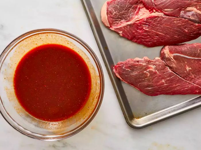 A steak on a baking sheet with a bowl of marinade beside it