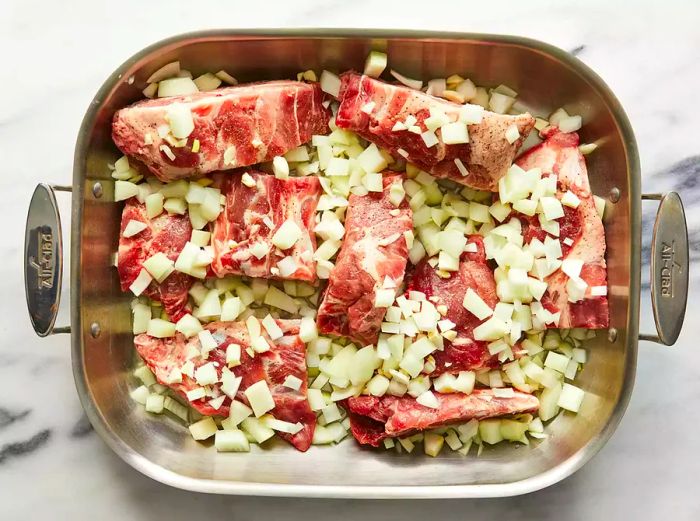 Bird's eye view of a roasting pan filled with raw, seasoned neck bones, topped with chopped onions and garlic.