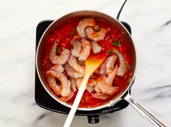 A top-down view of a pot with shrimp and tomato sauce being stirred to make shrimp Creole.