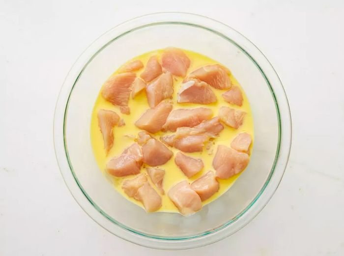 A large glass bowl filled with cubed chicken breast soaking in a rich egg and milk mixture.